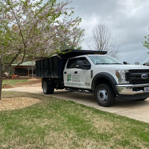 A white Canelo Landscape & Design truck parked on a concrete driveway with a residential area in the background. Landscaping equipment is visible in the distance near the house.