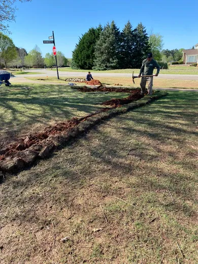 Image of the installation process of a french drain - a long trench has been dug through a grass yard. 