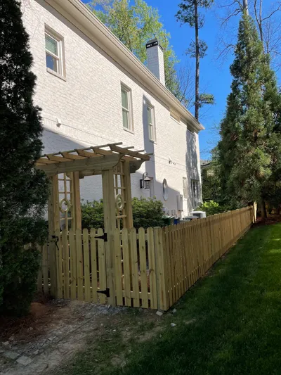Wood pergola with picket gate along fence line at a white brick home with mature trees