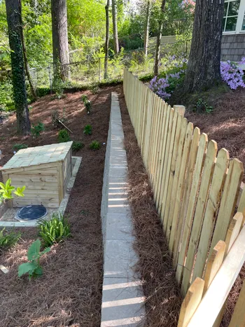 New wood privacy fence with stone walkway and pine straw mulch along a wooded backyard slope