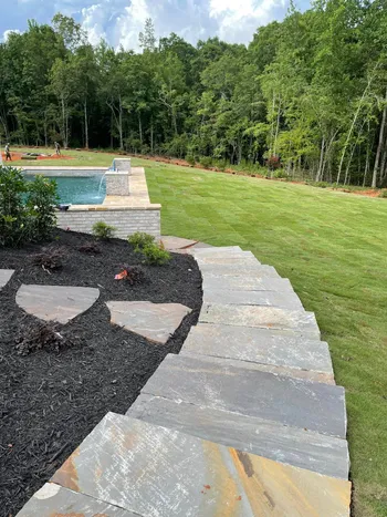 Large natural slate walkway and patio beside a new pool with fresh black mulch beds and stone retaining wall